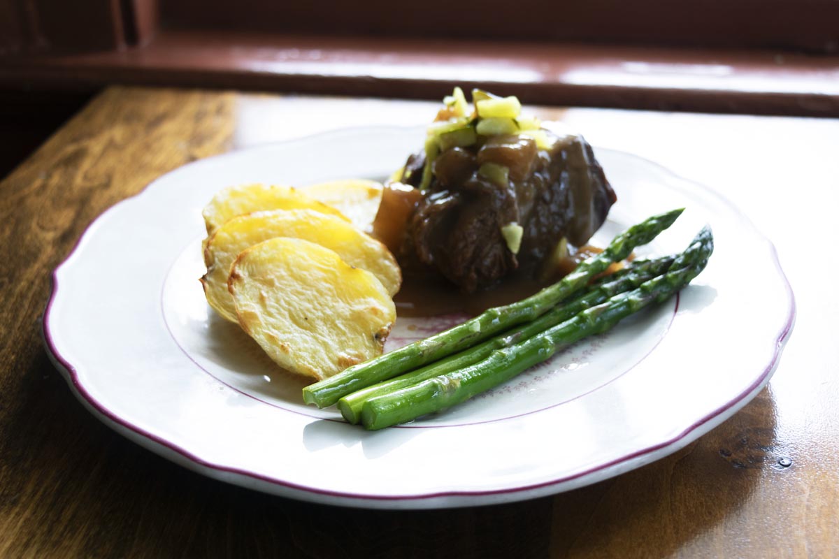 White plate filled with beef with gravy topped with chopped vegetables, scalloped potatoes, and asparagus, sitting on wooden table