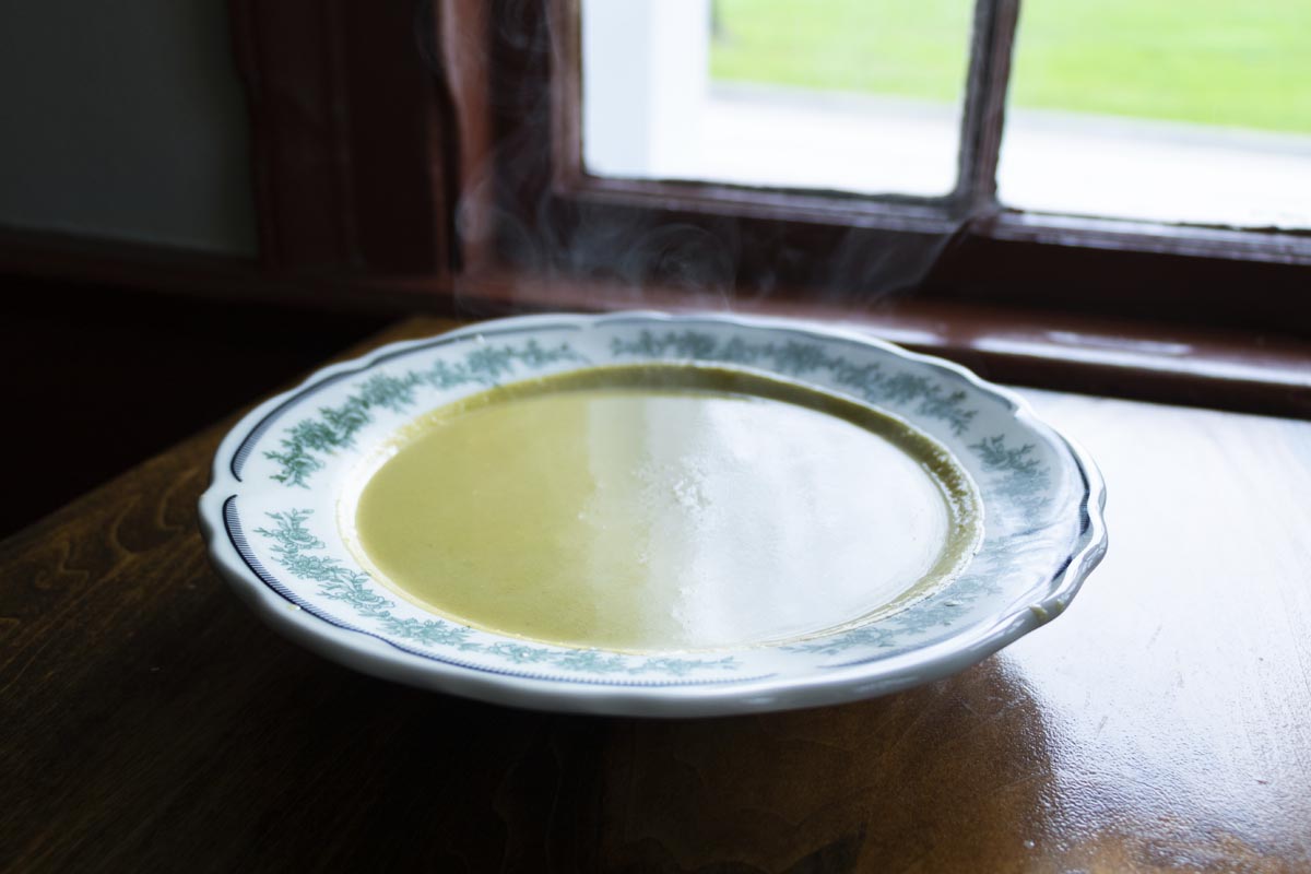 Shallow white bowl with green floral pattern filled with steaming pea-colored soup, sitting on wooden table in front of window