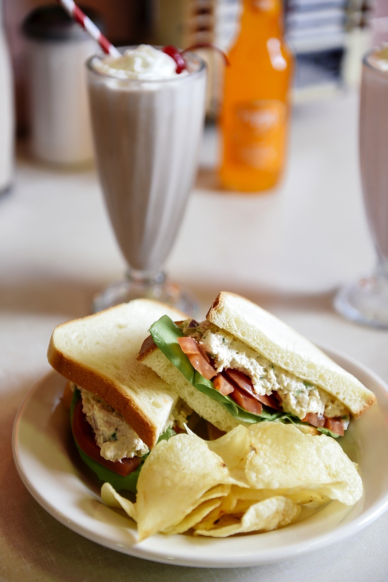 Plate with sandwich and potato chips sits on a counter along with a milkshake, orange Faygo in a bottle, and a napkin dispenser