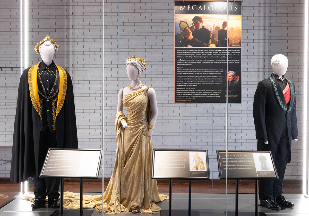 Two mannequins wearing suits and one in a gold dress, inside a glass display case