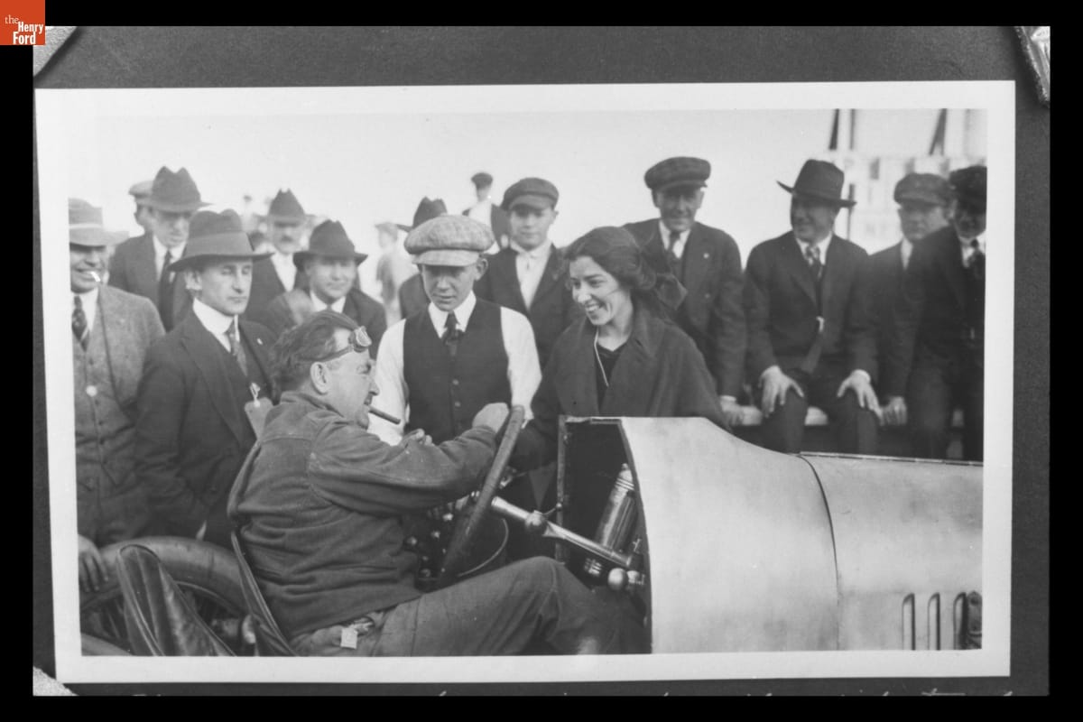 Crowd of men and one woman gathered around man with cigar sitting in open race car