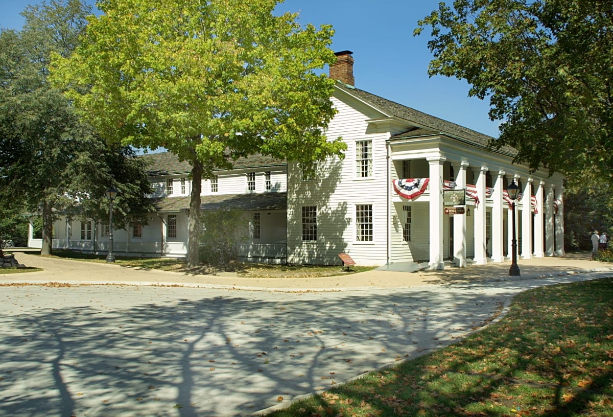 Large, two-story white wooden building with columns and second-floor balcony, among trees