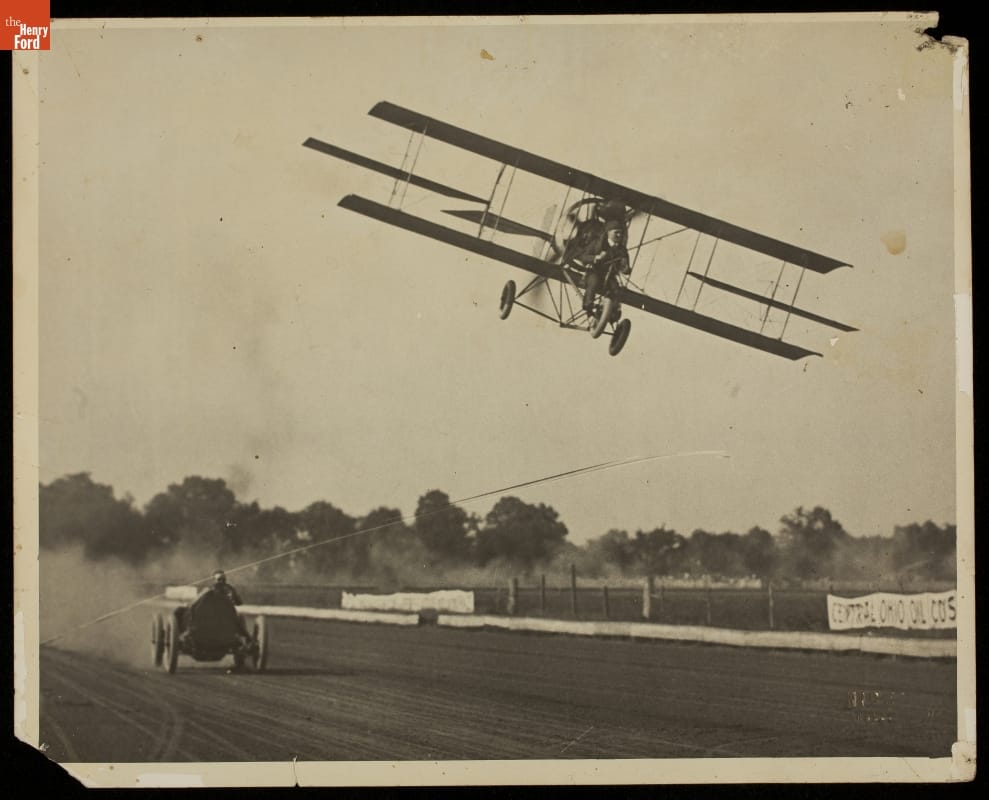 Car races on dirt racetrack while plane flies low overhead