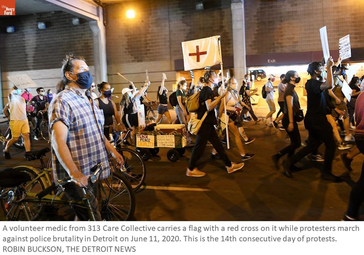 Detroit News Photograph of Volunteer Medic at Police Brutality Protest, Detroit, Michigan, June 11, 2020 Photo of people in protest march, some walking bicycles or holding signs; person in center carries a white flag with a red cross; also contains text